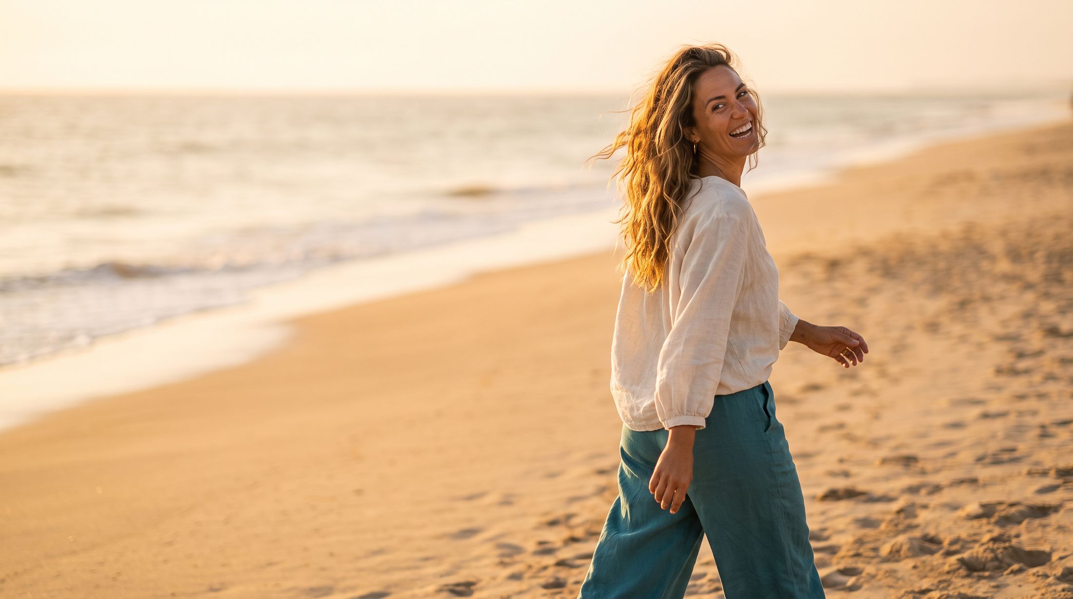 A woman in a linen blouse and teal trousers laughing as she looks back over her shoulder on a beach at golden hour.